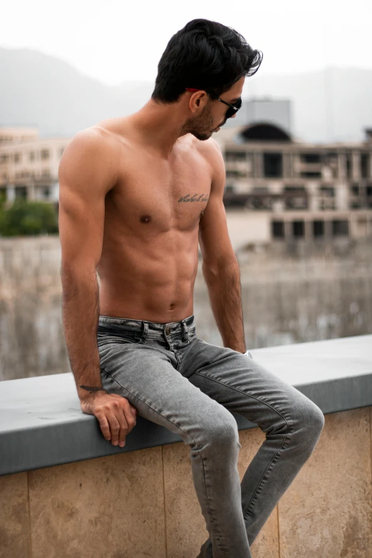 a shirtless man sitting on a ledge next to a body of water, by Cosmo Alexander, trending on pexels, renaissance, tight denim jeans, dressed in a gray, middle eastern, non binary model