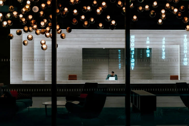 a person sitting at a table in front of a window, inspired by Cerith Wyn Evans, unsplash contest winner, intricate lights, infinite library, atrium, at night