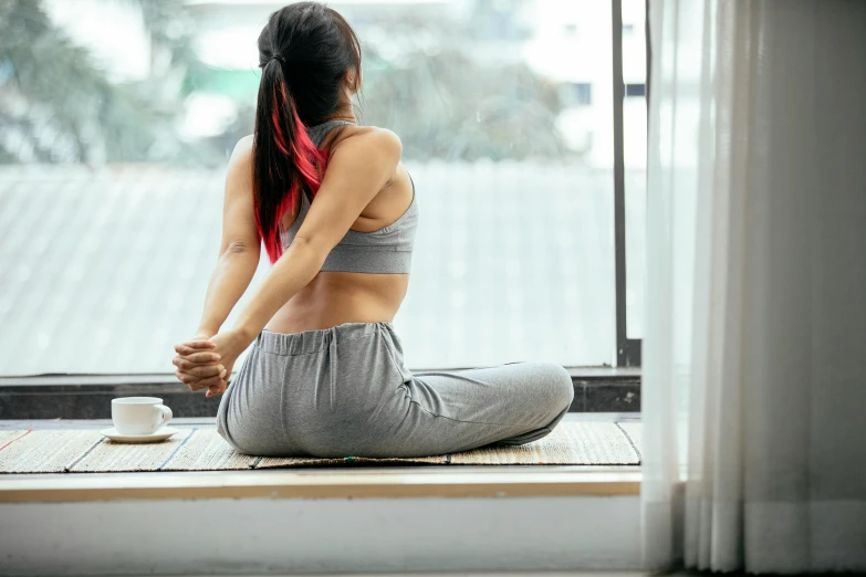 a woman sitting on a mat in front of a window, waist reaching ponytail, manuka, physical : tinyest midriff ever, morning time