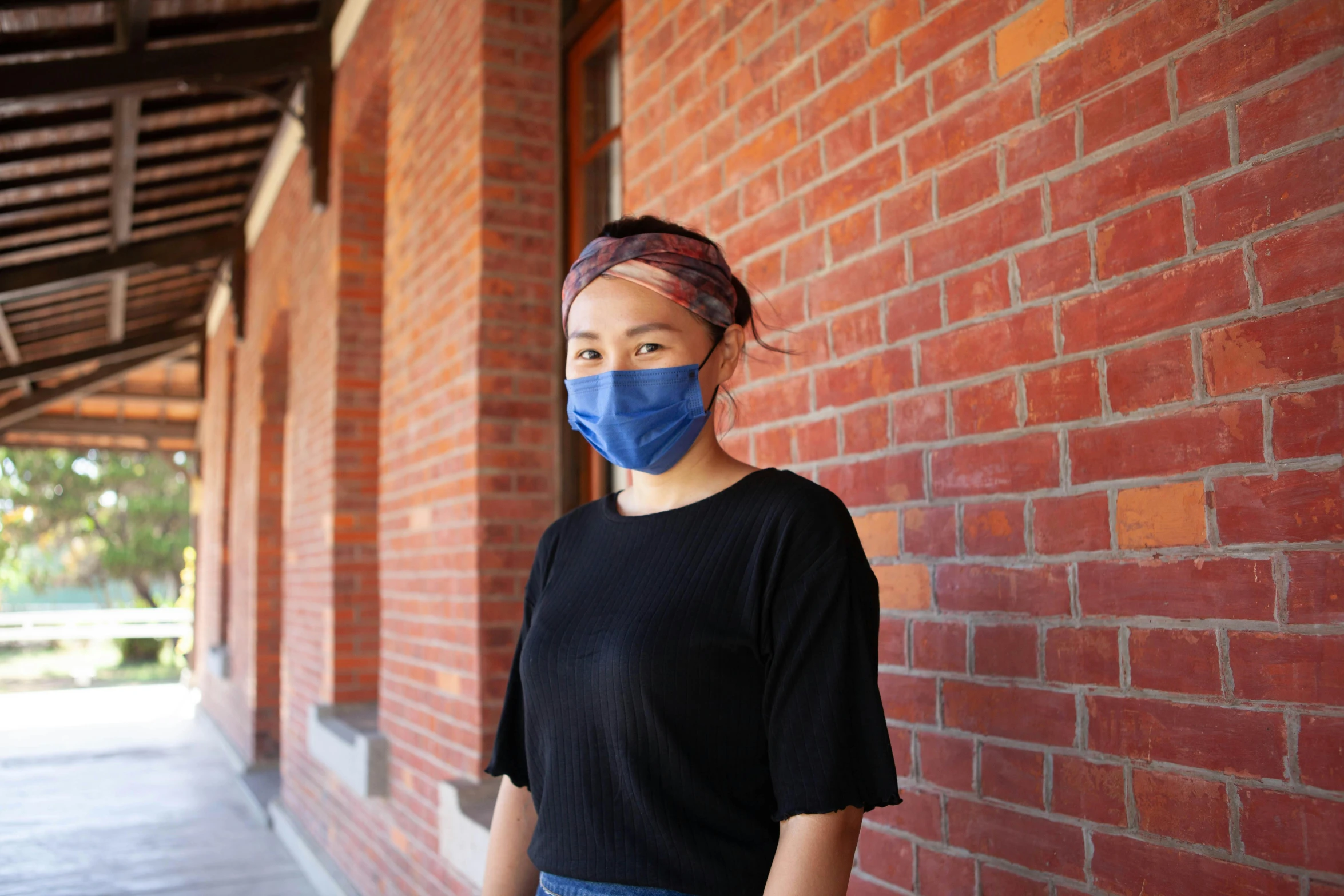 a woman standing in front of a brick wall wearing a face mask, inspired by Feng Zhu, blue, university, looking towards the camera, wearing facemask