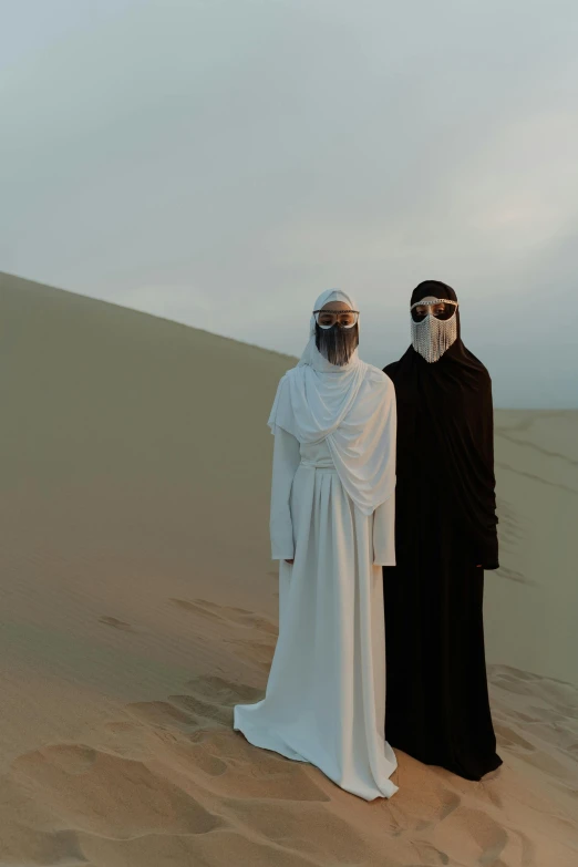 a couple of people that are standing in the sand, an album cover, by Ahmed Yacoubi, trending on unsplash, afrofuturism, wearing simple robes, masked, ☁🌪🌙👩🏾, 2 0 1 0 s