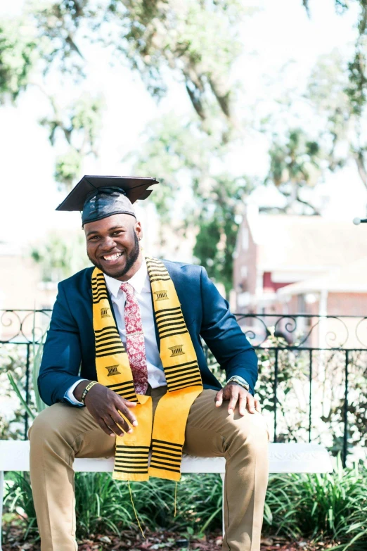 a man in a graduation cap and gown sitting on a bench, a portrait, by Carey Morris, unsplash, yellow navy teal black and gold, brown skinned, christian, sitting down