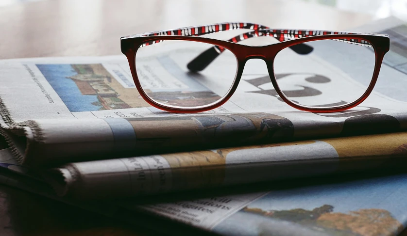 a pair of glasses sitting on top of a stack of newspapers, a picture, unsplash, private press, wearing red tainted glasses, square rimmed glasses, up close picture, on a wooden table