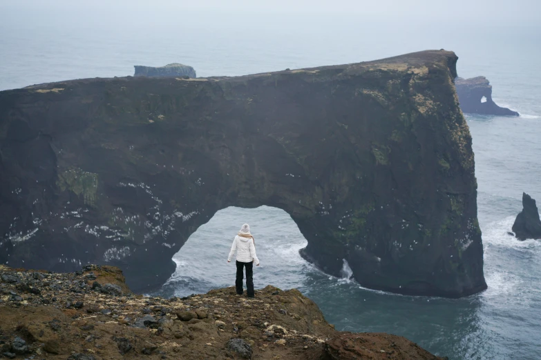a person standing on top of a cliff next to the ocean, by Hallsteinn Sigurðsson, happening, massive arch, view from the distance, photographed for reuters, photograph taken in 2 0 2 0