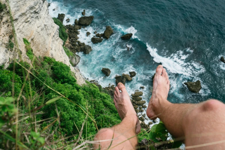 a man sitting on top of a cliff next to the ocean, pexels contest winner, hairy legs, bali, vertigo - inducing, happy toes