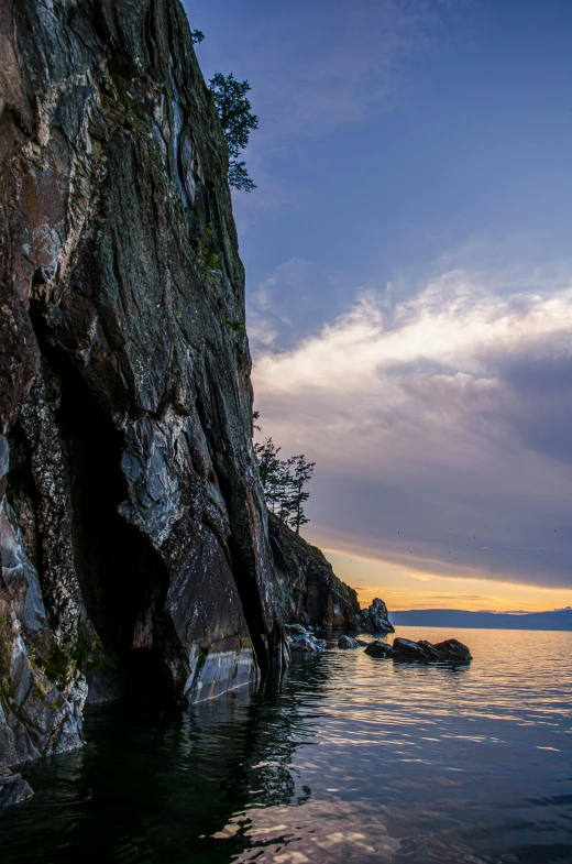 a cave in the middle of a body of water, by Doug Ohlson, lake baikal in the background, sunset south, rock wall, lush surroundings