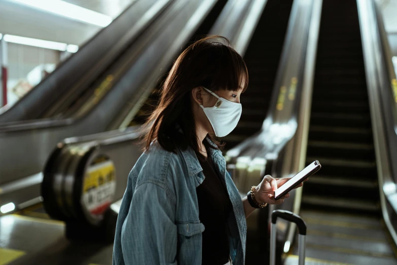 a woman wearing a face mask standing next to an escalator, pexels contest winner, she is holding a smartphone, square, scientific, underground scene