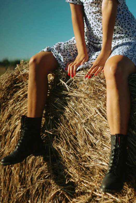 a woman sitting on top of a bale of hay, trending on pexels, black leather boots, shows a leg, summer evening, ad image