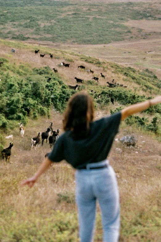 a woman standing on top of a lush green hillside, by Lucia Peka, trending on unsplash, happening, many goats, waving arms, grainy film still, friends