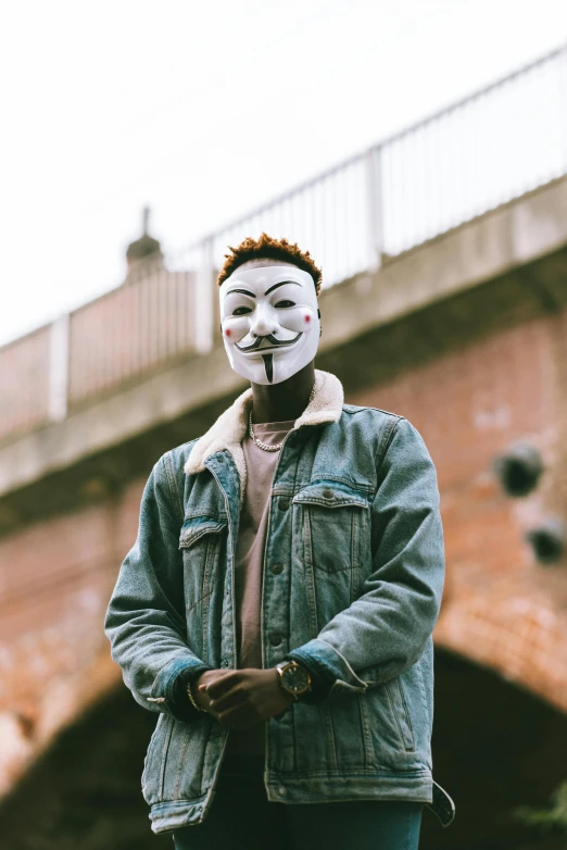 a man wearing a mask standing in front of a bridge, trending on pexels, graffiti, donald glover, non-binary, anonymous as a sausage, bank robbery movies