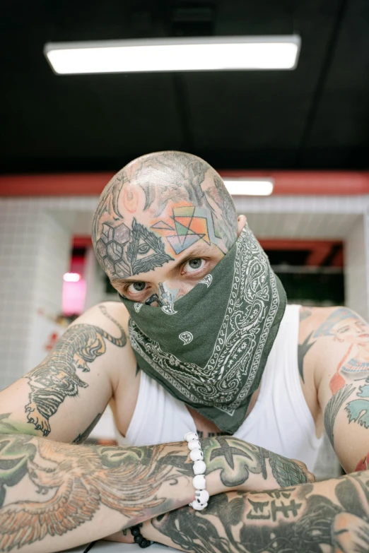 a man with tattoos sitting at a table, a tattoo, by Brian 'Chippy' Dugan, trending on pexels, lowbrow, black bandana mask, centered shoulders up view, 2000s photo, mysterio