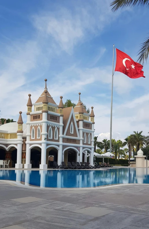 a large building with a flag flying in front of it, inspired by Nevin Çokay, hurufiyya, tropical pool, turrets, holiday resort, exterior photo