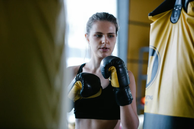 a woman standing next to a punching bag, by Emma Andijewska, pexels contest winner, happening, black and yellow tracksuit, avatar image, manuka, 1614572159