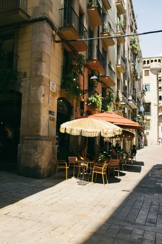 a sidewalk with tables and umbrellas in front of a building, by Carlo Carrà, unsplash, gothic quarter, gif, sunlit, panoramic