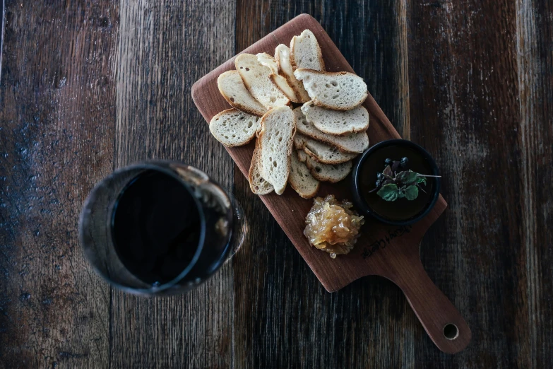 a wooden cutting board topped with bread next to a glass of wine, black on black, birdseye view, local foods, background image