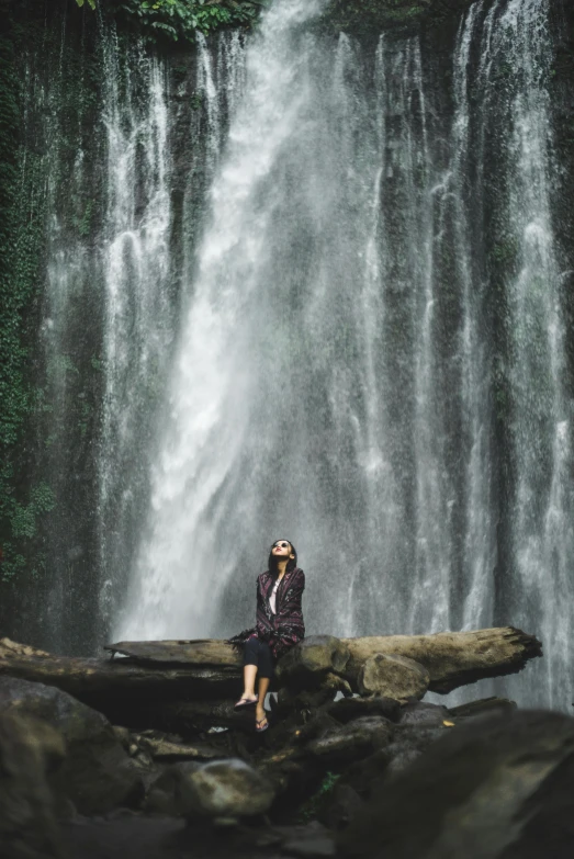 a woman sitting on a log in front of a waterfall, by Julia Pishtar, sumatraism, high-quality photo, trending photo