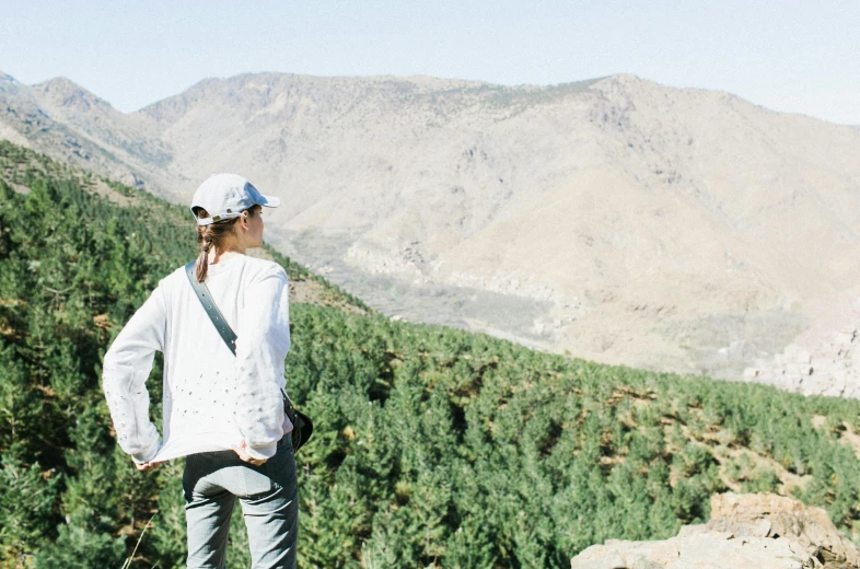 a woman standing on top of a lush green hillside, by Alice Mason, trending on unsplash, les nabis, morocco, pine trees in the background, outlive streetwear collection, looking at the mountains