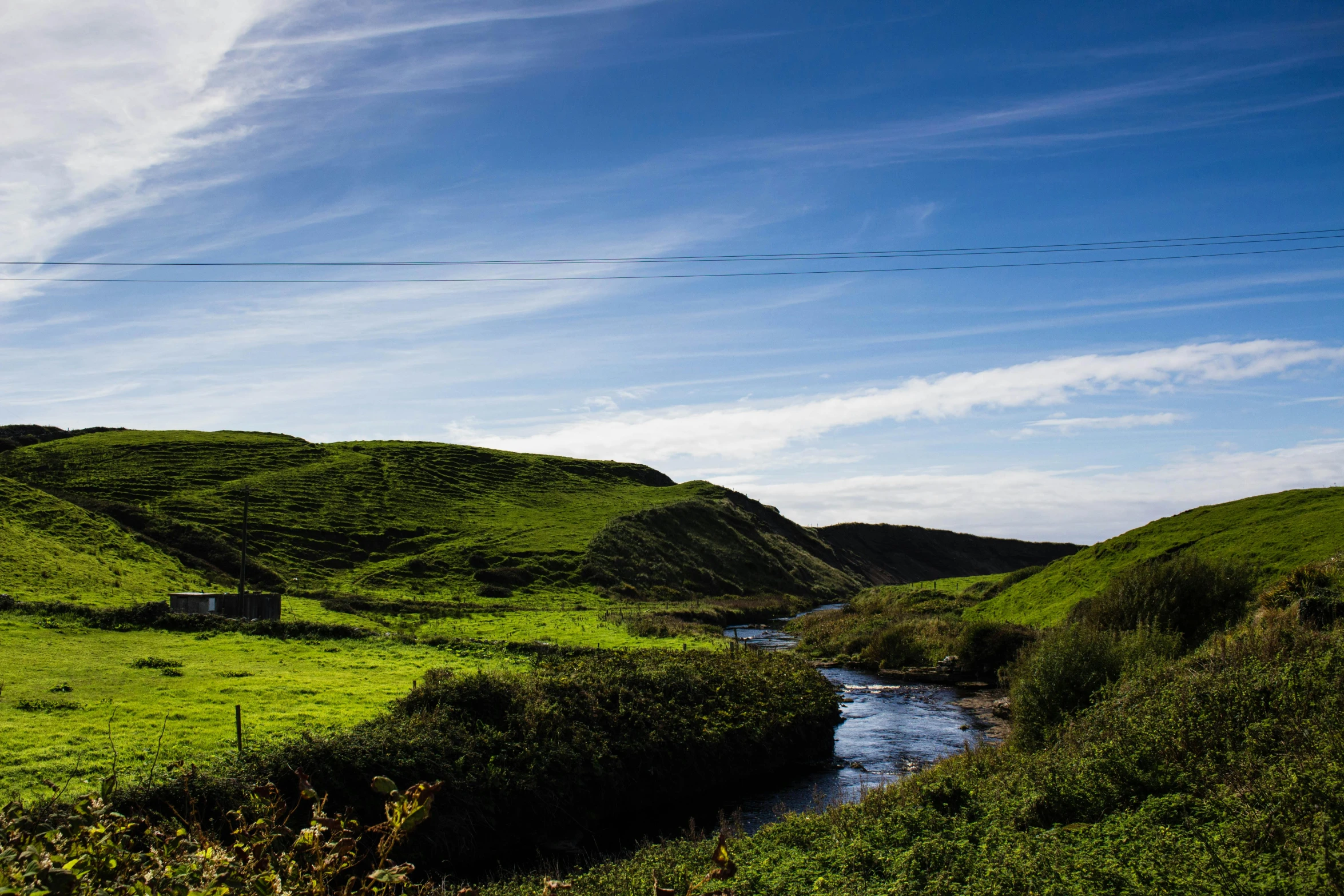 a river running through a lush green hillside, by Andrew Allan, unsplash, hurufiyya, blue sky and green grassland, near farm, coastal, marsden