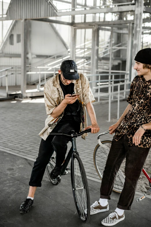 a man riding a bike next to a woman on a bike, an album cover, by Nina Hamnett, trending on pexels, skatepark, looking at his phone, two young men, brown