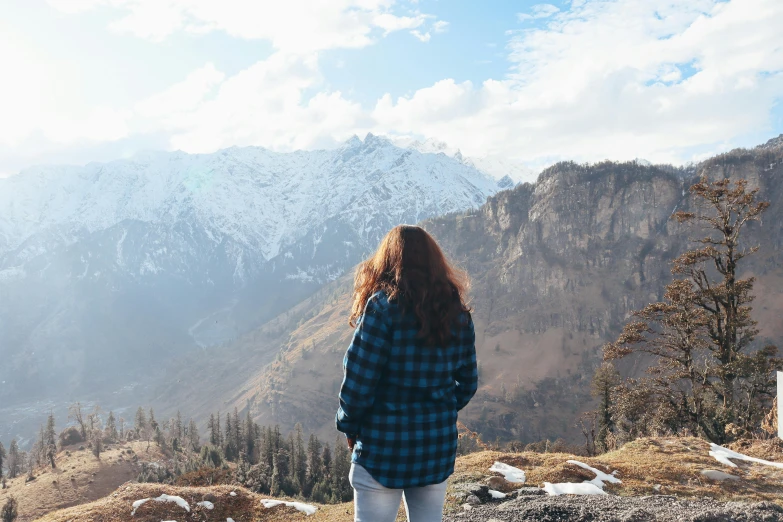 a woman standing on top of a rocky hill, a picture, trending on pexels, wearing a flannel shirt, majestic snowy mountains, background image, young himalayan woman