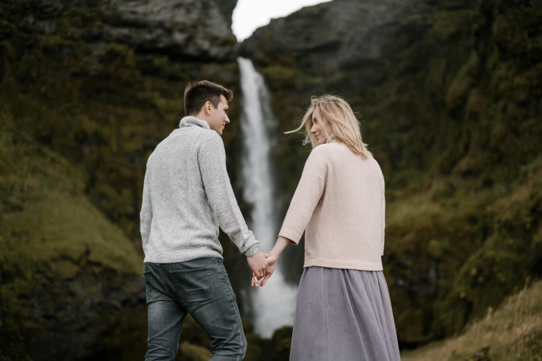 a man and woman holding hands in front of a waterfall, by Hallsteinn Sigurðsson, pexels contest winner, hurufiyya, attractive girl, looking from behind, grey, romantic lead