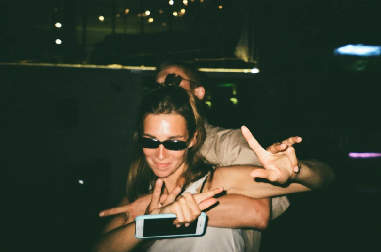 a woman standing next to a man holding a cell phone, a polaroid photo, by Niko Henrichon, unsplash, having a cool party, giving the middle finger, dark shades, sunfaded