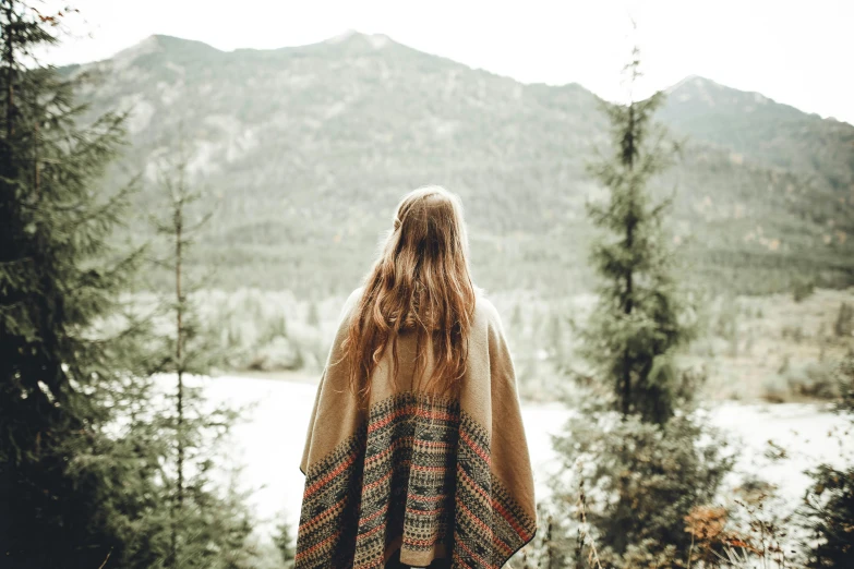 a woman standing in front of a lake wrapped in a blanket, trending on pexels, visual art, brown colored long hair, overlooking a valley with trees, slightly pixelated, poncho
