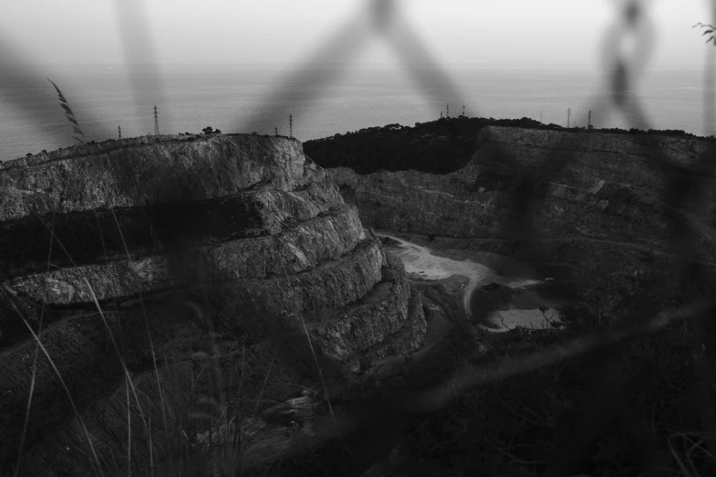 a view of the ocean through a chain link fence, a black and white photo, by Mathias Kollros, pexels contest winner, surrealism, in dusty open pit mine, cliff side at dusk, agrigento, ( apocalyptic ) 8 k