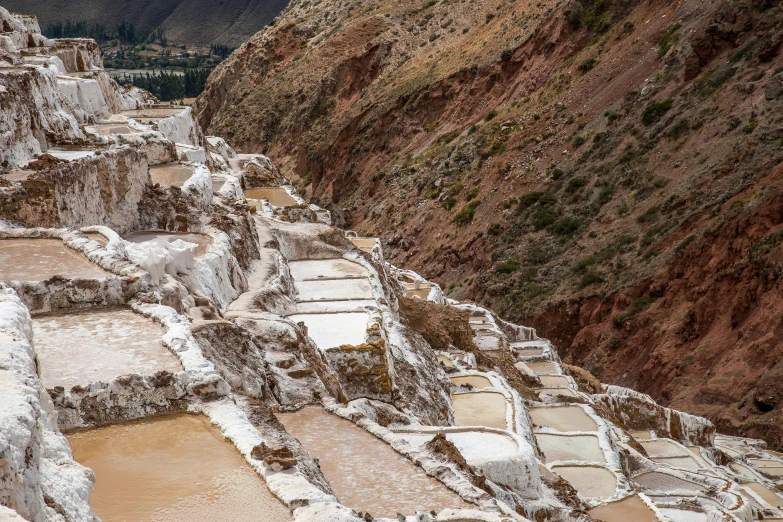 a group of salt pans sitting on the side of a mountain, a mosaic, by Julia Pishtar, trending on unsplash, quechua, flowing white robes, rail tracks lead from the mine, pink water in a large bath