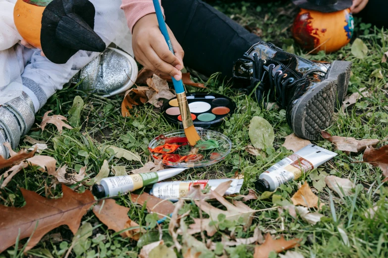 a group of people sitting on top of a grass covered field, a photorealistic painting, by Julia Pishtar, pexels contest winner, process art, holding a paintbrush in his hand, during autumn, facepaint, flatlay