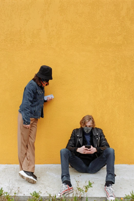 a couple of people sitting on the side of a building, an album cover, trending on pexels, yellow walls, bank robbery, looking at his phone, before a stucco wall