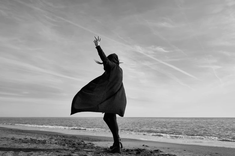 a woman standing on top of a beach next to the ocean, a black and white photo, by Caro Niederer, pexels contest winner, conceptual art, waving robe movement, wearing black coat, superhero, 15081959 21121991 01012000 4k