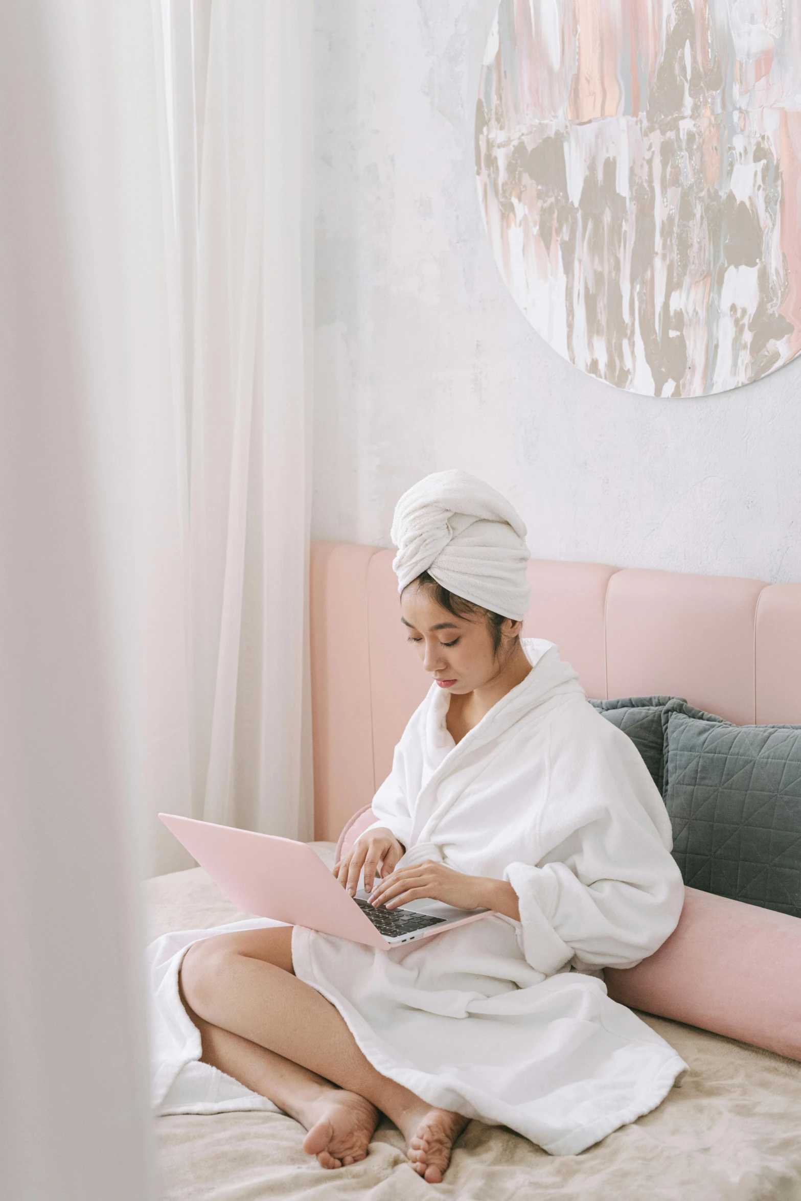 a woman sitting on a bed using a laptop, by Julia Pishtar, pexels contest winner, happening, pastel pink robes, skincare, white sarong, hair styled in a bun