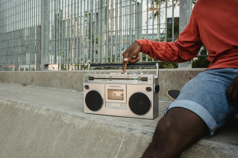 a man sitting on a ledge next to a boombox, brown, connectivity, holding it out to the camera, on the concrete ground