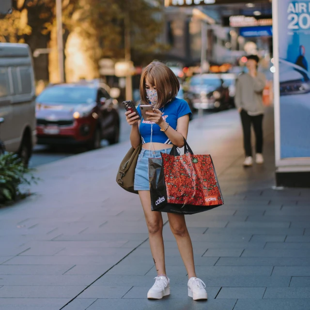 a woman standing on a sidewalk looking at her phone, pexels contest winner, bra and shorts streetwear, in australia, malls, people are wearing masks