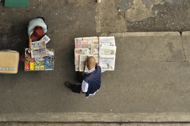 a man sitting on the sidewalk reading a newspaper, by Matt Stewart, pexels contest winner, private press, aerial view from above, in sao paulo, stack of comics on the floor, view from a news truck