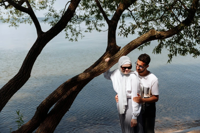 a man and a woman standing next to a tree, by Nazmi Ziya Güran, pexels contest winner, hurufiyya, white sarong, lakeside, holding a bottle of arak, hijab