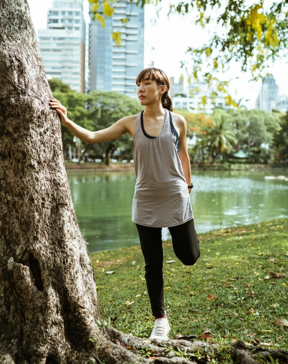 a woman standing next to a tree in a park, inspired by Ruth Jên, unsplash, happening, wearing fitness gear, singapore, grey, view