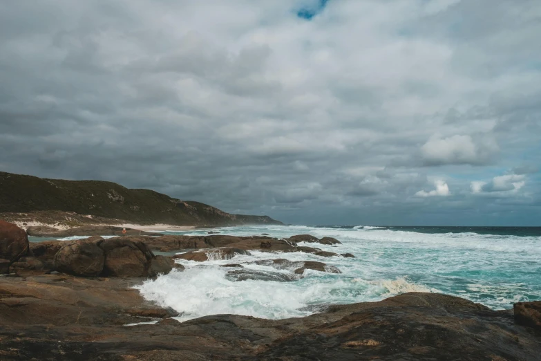 a man standing on top of a rocky beach next to the ocean, pexels contest winner, process art, south african coast, big overcast, cresting waves and seafoam, the emerald coast