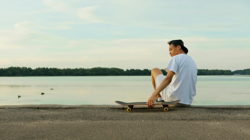 a man sitting on top of a skateboard next to a body of water, inspired by mads berg, pexels contest winner, realism, plain background, profile pic, serene environment, male teenager