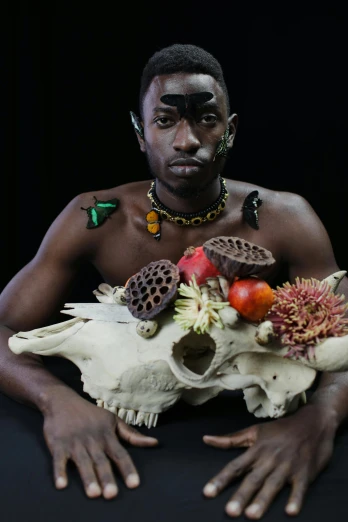 a man sitting at a table with a skull in front of him, an album cover, by Jessie Alexandra Dick, pexels contest winner, afrofuturism, made of flowers and fruit, black skin, model posing, a horned