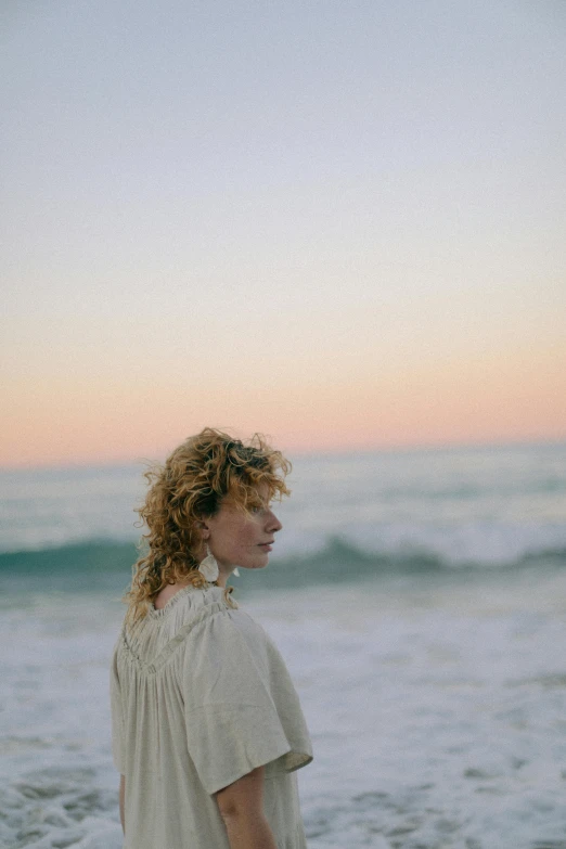 a woman standing on top of a beach next to the ocean, an album cover, unsplash, renaissance, pale skin curly blond hair, soft evening lighting, profile image, curly bangs