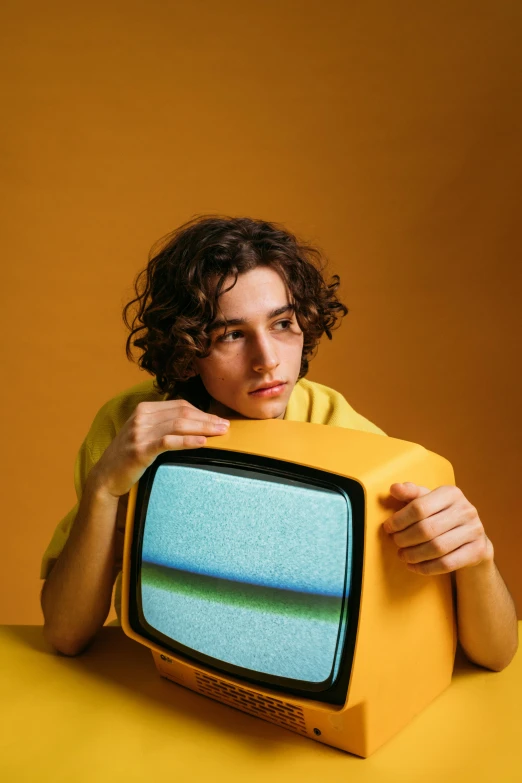 a woman holding a television in front of her face, an album cover, trending on pexels, portrait of timothee chalamet, wavy hair yellow theme, sitting down, high quality photo