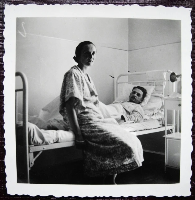 a woman sitting on a bed next to a man, a black and white photo, by Evaline Ness, at the hospital in patient gown, album cover, soviet - era, taken in the early 2020s