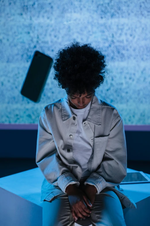 a woman sitting on a bench in front of a tv, afrofuturism, wearing a lab coat, projection mapping, sad prisoner holding ipad, curly afro