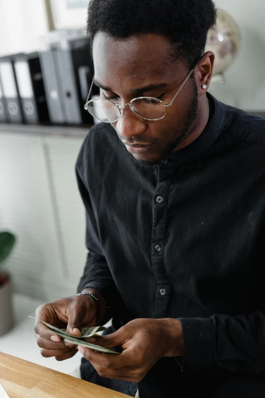 a man sitting at a desk looking at a cell phone, pexels contest winner, renaissance, black turtle neck shirt, ( ( dark skin ) ), man with glasses, cash on a sidetable