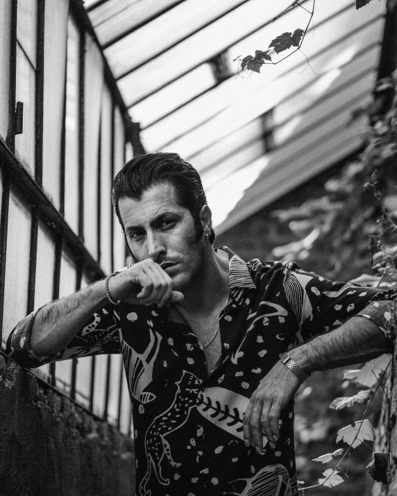 a black and white photo of a man leaning against a wall, rockabilly hair, adam sandler, mateus 9 5, amongst foliage