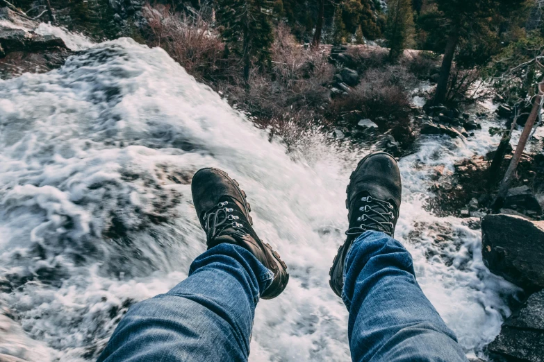 a person standing on top of a rock next to a river, jeans and boots, 8 feet fall, pov photo, white water