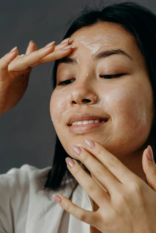 a woman is rubbing her face with her hands, trending on pexels, renaissance, asian face, cream dripping on face, goggles on forehead, on a gray background