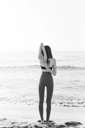 a woman standing on top of a beach next to the ocean, a black and white photo, unsplash, arabesque, wearing fitness gear, back - view, character posing, wearing a crop top